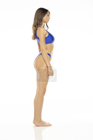 Foto de Joven mujer morena hermosa en traje de baño bikini azul posando sobre un fondo de estudio blanco. Vista lateral. - Imagen libre de derechos