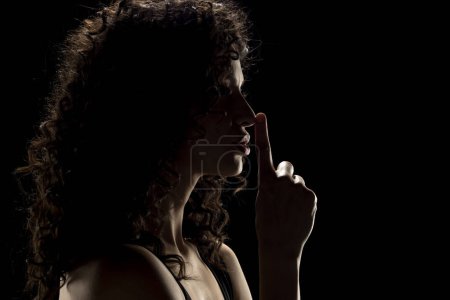 Foto de Silueta de mujer desconocida con la cara en la sombra sosteniendo el dedo en sus labios sobre un fondo negro de estudio - Imagen libre de derechos