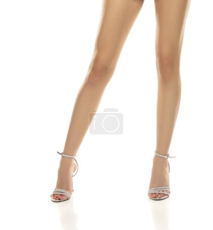 Foto de Acercamiento de las piernas bonitas de una mujer en un vestido blanco corto y sandalias de tacón alto sobre un fondo de estudio blanco. - Imagen libre de derechos