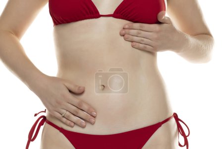 Foto de Mujer joven en bikini rojo aplica producto cosmético en su vientre sobre fondo de estudio blanco. - Imagen libre de derechos