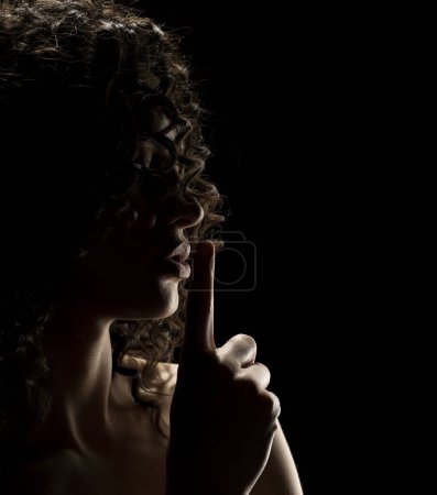 Foto de Silueta de mujer desconocida con la cara en la sombra sosteniendo el dedo en sus labios sobre un fondo negro de estudio - Imagen libre de derechos