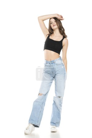 Téléchargez les photos : Jeune mannequin femme portant un jean déchiré et une chemise noire sans manches posant sur un fond de studio blanc. Vue de face - en image libre de droit