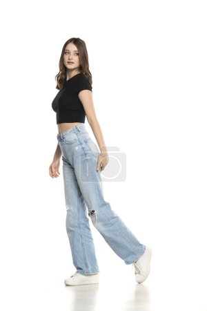 Photo for Young female model wearing ripped jeans and black shirt walking on a white studio background. Side, profilet view - Royalty Free Image