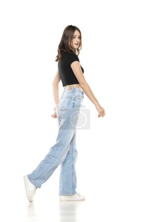 Foto de Joven modelo femenina con pantalones vaqueros rasgados y camisa negra caminando sobre un fondo de estudio blanco. Lado, vista de perfil - Imagen libre de derechos