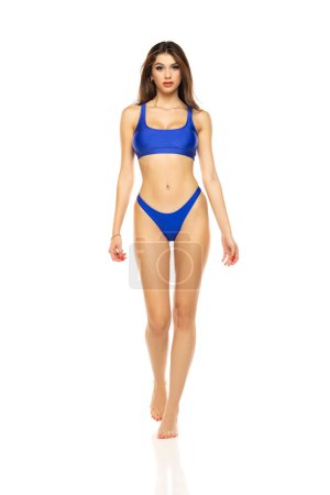 Jeune belle femme brune en maillot de bain bikini bleu marchant sur un fond de studio blanc.