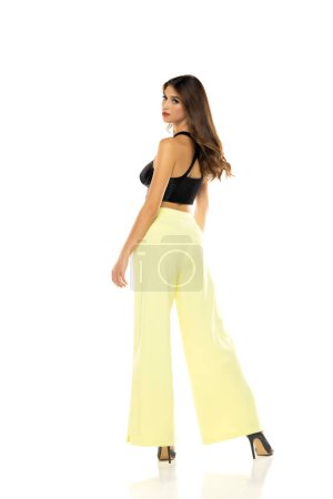 Photo for Fashion model posing in black blouse, yellow loose pants and high heel shoes. Rear view on a white studio background - Royalty Free Image