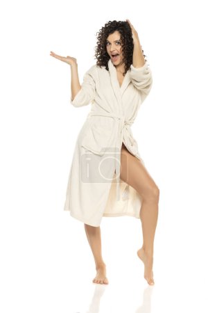 Téléchargez les photos : Jeune femme heureuse posant en peignoir et tenant un objet imaginaire sur un fond de studio blanc - en image libre de droit