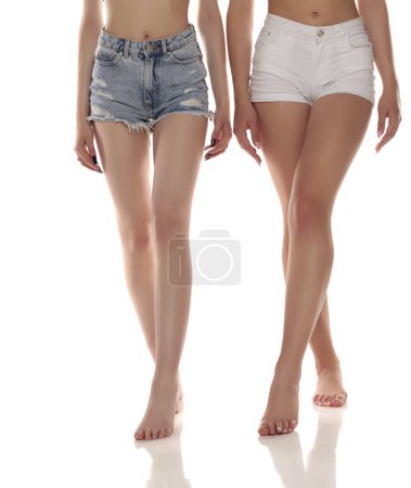 Foto de Fotografía de cerca de dos mujeres descalzas con pantalones vaqueros cortos sobre fondo blanco del estudio. Vista frontal. - Imagen libre de derechos