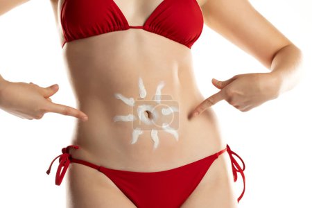 Téléchargez les photos : Jeune femme en bikini rouge avec des produits cosmétiques en forme de soleil sur le ventre sur fond de studio blanc. - en image libre de droit