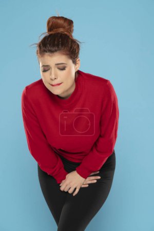 Téléchargez les photos : Femme rousse souffrant de douleur et tenant ses mains dans le bas-ventre sur un fond bleu studio - en image libre de droit