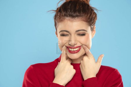 Téléchargez les photos : Jeune rousse femme forçant son sourire avec ses doigts sur fond bleu studio - en image libre de droit