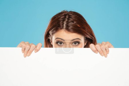 Photo for Banner of young unhappy redhead woman feeling sad and peeking behind empty white canvas frame for text or advertising isolated on blue studio background - Royalty Free Image