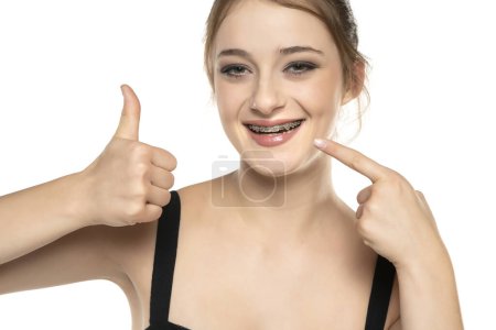 Photo for Cheerful happy young woman with blonde hair gesturing thumb up while pointing finger at braces on her teeth isolated over white studio background. The concept of a healthy snow-white smile. Daily dental care - Royalty Free Image