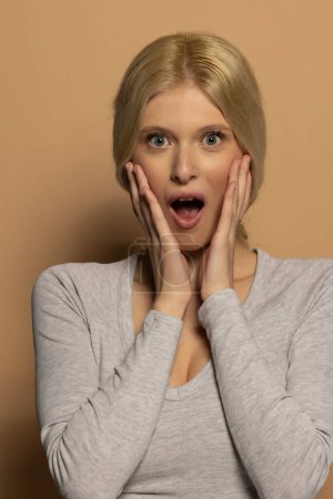 Téléchargez les photos : Portrait de belle jeune femme choquée aux longs cheveux blonds attachés sur fond studio beige - en image libre de droit