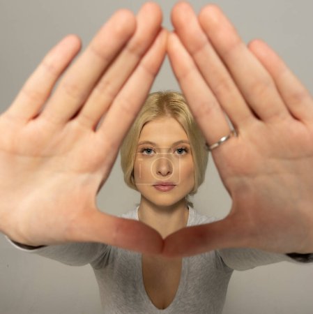 Foto de Atractiva joven mujer mirando a través de los dedos marco en fondo gris estudio - Imagen libre de derechos