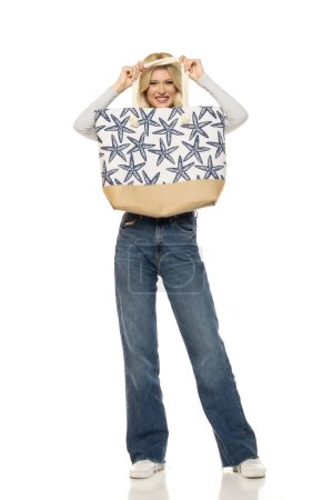 Foto de Hermosa mujer rubia feliz joven en jeans con bolso de playa en fondo de estudio blanco - Imagen libre de derechos
