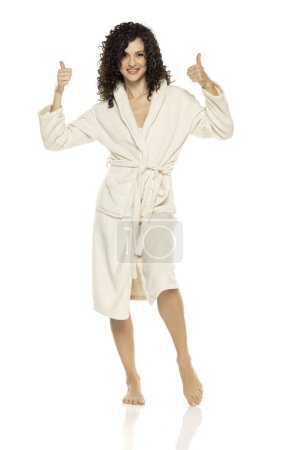 Photo for Young happy woman posing in bathrobe and showing thumbs up on a white studio background - Royalty Free Image