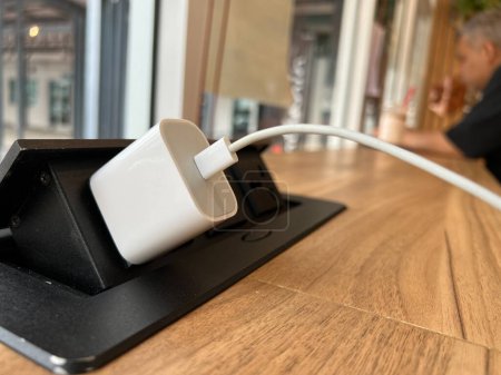 Close up the white charger connected to the pop-up electrical outlet on the wooden table in the coffee shop