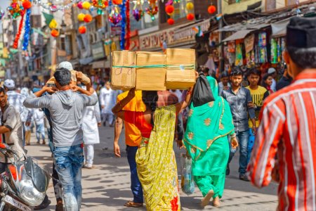 Foto de AJMER, INDIA - MARCH 3 2018: Colorful scene of woman carries three boxes on her head by the people on the Dargah Bazar road. - Imagen libre de derechos