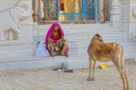 Foto de PUSHKAR, INDIA - MARCH 3 2018: Colorful scene of a shoeless woman in traditional clothes resting on the temple steps. - Imagen libre de derechos