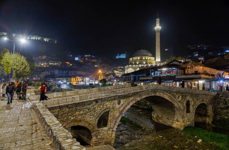 Téléchargez les photos : PRIZREN, KOSOVO - 12 NOVEMBRE 2022 : Les gens marchent par le vieux pont en pierre, la mosquée ottomane Sinan Pacha et la rivière Bistrica dans le centre-ville pendant la froide nuit de novembre. longue exposition - en image libre de droit