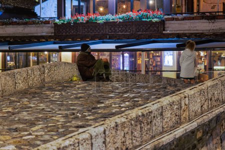 Photo for PRIZREN, KOSOVO - NOVEMBER 12, 2022: A lonely man sitting and begging on an old bridge in city center by night. long exposure - Royalty Free Image