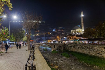 Téléchargez les photos : PRIZREN, KOSOVO - 12 NOVEMBRE 2022 : Les gens marchent par le vieux pont en pierre, la mosquée ottomane Sinan Pacha et la rivière Bistrica dans le centre-ville pendant la froide nuit de novembre. longue exposition - en image libre de droit