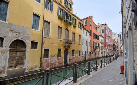 Foto de Una pintoresca vista de un canal de Venecia, con una pasarela de piedra, barandilla de hierro forjado, y un puente de piedra distante. - Imagen libre de derechos