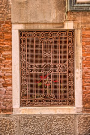 Photo for Antique window with metal grilles on a Venetian house, with a hint of a vibrant red flower, captured using a tilt shift lens emphasizing a narrow vertical focus. - Royalty Free Image