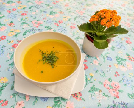 Téléchargez les photos : Riche soupe de courge butternut jaune garnie de persil à côté d'un vase de fleurs orange sur une nappe à motifs floraux. - en image libre de droit