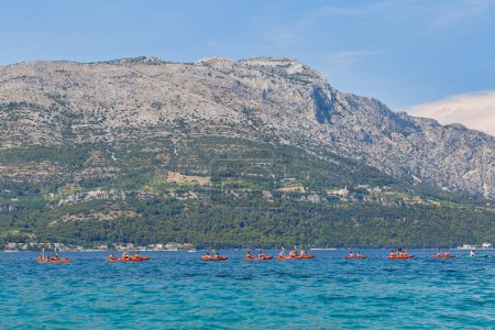 Photo for KORCULA, CROATIA - June 24, 2019: A group of kayaks with tourists glide on the water with the Peljesac peninsula and Mount St. Ilija in the backdrop. - Royalty Free Image