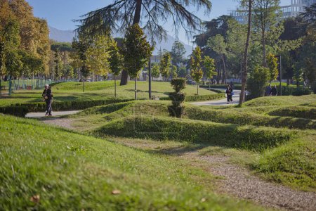 Foto de TIRANA, ALBANIA - 20 de octubre de 2019: Parque urbano vibrante con exuberante vegetación, colinas artificiales y peatones paseando. - Imagen libre de derechos