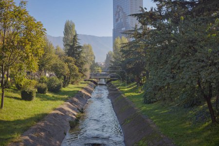 Foto de TIRANA, ALBANIA - 20 de octubre de 2019: Corriente que fluye a través del canal de las ciudades rodeada de vegetación con edificios modernos en el fondo. - Imagen libre de derechos