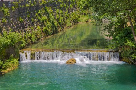 Foto de Una vista impresionante del río Pliva en cascada que fluye serenamente a través de la pintoresca e histórica ciudad de Jajce, rodeada de exuberante vegetación y una vibrante atmósfera de verano. - Imagen libre de derechos