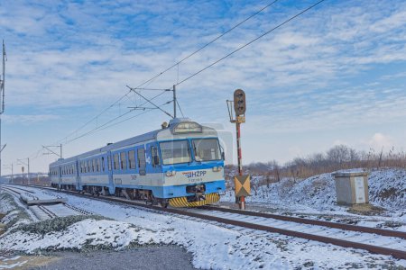 Foto de DUGO SELO, CROACIA - 25 de enero de 2023: Un tren azul de pasajeros HZPP se acerca a la estación en un día de invierno nevado, con el paisaje rural de Dugo Selo bajo un cielo parcialmente nublado. - Imagen libre de derechos