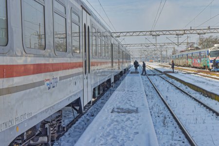 Foto de DUGO SELO, CROACIA - 25 de enero de 2023: Pasajeros caminando junto a un tren estacionario en las vías cubiertas de nieve en la estación de tren de Dugo Selo en un día despejado de invierno. - Imagen libre de derechos