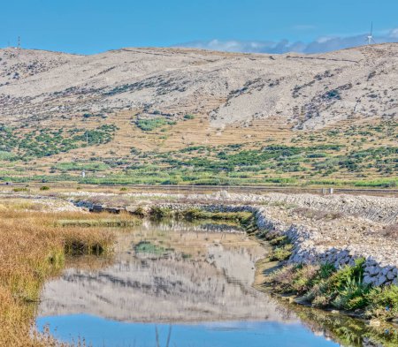 Téléchargez les photos : Paysage panoramique avec un canal d'eau au milieu du terrain aride de l'île de Pag, sous un ciel dégagé. - en image libre de droit