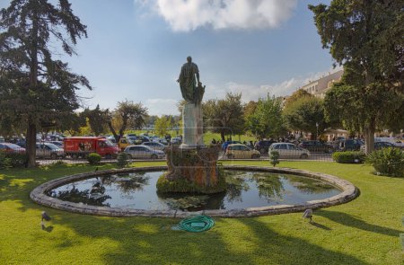 Foto de CORFU, GRECIA - 21 de octubre de 2022: Estatua de Sir Frederick Adam del escultor Pavlos Prosalentis, situada contra un estanque reflectante en medio de la vegetación de un parque de la ciudad, con un animado telón de fondo. - Imagen libre de derechos