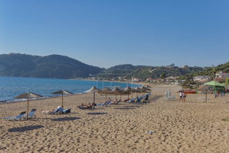 Foto de CORFU, GRECIA - 21 de octubre de 2022: Los visitantes se relajan en las tumbonas bajo sombrillas de paja en una playa de arena en Corfú, con las exuberantes colinas verdes en el fondo. - Imagen libre de derechos