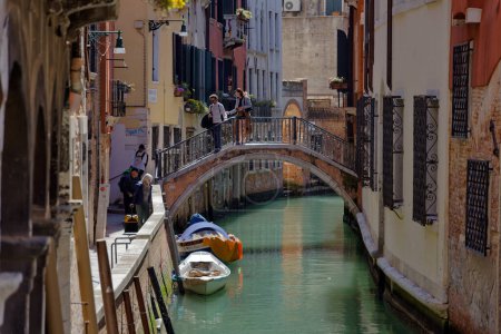 Foto de VENECIA, ITALIA - 10 DE ABRIL DE 2023: Gente cruzando un pequeño puente de arco en una calle del canal, encarnando el encanto del casco antiguo por excelencia. - Imagen libre de derechos