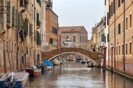 Foto de VENECIA, ITALIA - 11 ABRIL 2023: Pequeño puente de arco de cruce en una calle del canal, encarnando el encanto del casco antiguo por excelencia. - Imagen libre de derechos
