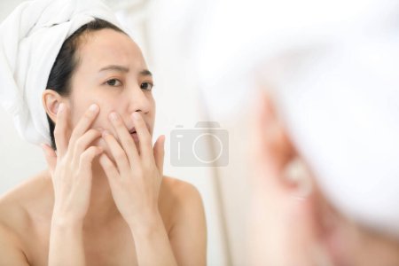 Problema de piel. Preocupado jóvenes mujeres asiáticas haciendo estallar espinilla en la mejilla, mientras que de pie cerca del espejo en el baño. Chica infeliz inspeccionando la cara, sufriendo acné, se centran en la reflexión