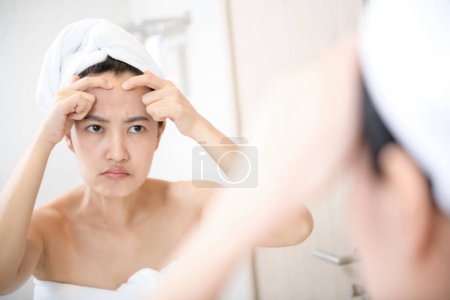 Téléchargez les photos : Problème de peau. Concerné jeunes femmes asiatiques qui éclatent bouton sur la joue tout en se tenant près du miroir dans la salle de bain. Fille malheureuse inspectant le visage, l'acné souffrant, se concentrer sur la réflexion - en image libre de droit