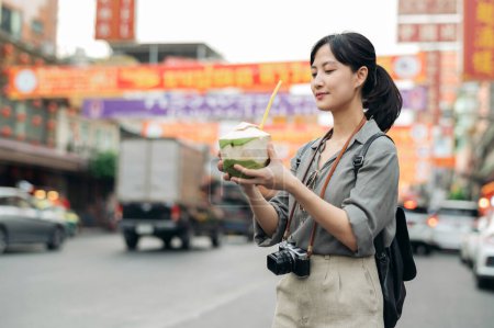 Téléchargez les photos : Heureux jeune voyageur asiatique sac à dos boire un jus de noix de coco au marché de la nourriture de rue de la ville de Chine à Bangkok, Thaïlande. - en image libre de droit