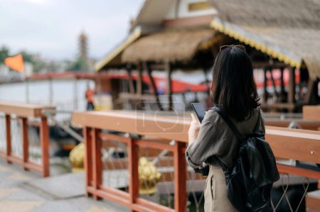 Téléchargez les photos : Young Asian woman backpack traveler using mobile phone in express boat pier on Chao Phraya River in Bangkok. - en image libre de droit