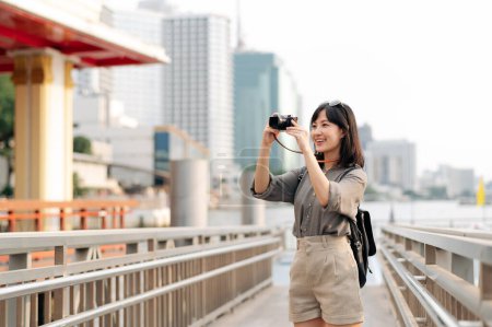 Young Asian woman backpack traveler using a camera in express boat pier on Chao Phraya River in Bangkok. Traveler checking out side streets.