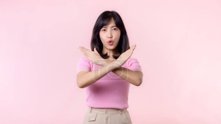 Téléchargez les photos : Portrait jeune femme asiatique sérieuse avec geste transversal montrant stop, non, faux, déni, signe de rejet isolé sur fond de studio pastel rose. nier et concept de symbole d'expression négative. - en image libre de droit