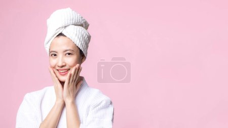 Foto de Hermosa mujer asiática joven feliz con la piel limpia de la cara en toalla y albornoz, traje de spa sobre fondo rosa. Cuidado de la piel, tratamiento, terapia de bienestar, cuidado facial, salud femenina de belleza, concepto de cosmetología - Imagen libre de derechos