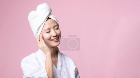 Foto de Joven hermosa mujer asiática feliz con la piel limpia de la cara en toalla y albornoz, traje de spa sobre fondo rosa. Cuidado de la piel, tratamiento, terapia de bienestar, cuidado facial, salud femenina de belleza, concepto de cosmetología - Imagen libre de derechos
