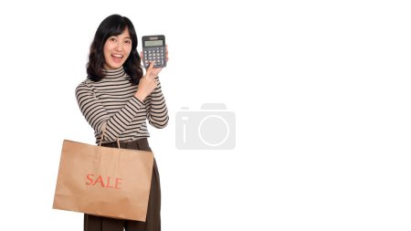 Téléchargez les photos : Jeune femme asiatique uniforme décontracté tenant sac de papier shopping et calculatrice sur fond blanc. Shopping concept de réduction. - en image libre de droit